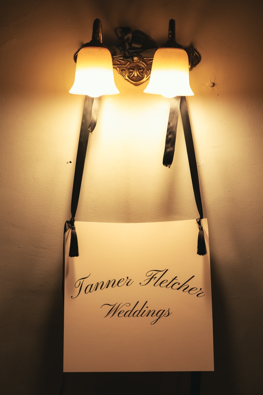 Tanner Fletcher Weddings sign with elegant black script lettering suspended by black satin ribbons beneath a warm wall sconce during Tanner Fletcher's immersive bridal fashion show at New York Bridal Fashion Week.